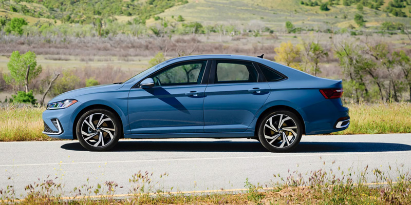 A blue sedan parked on the side of the road with a green, hilly background.
