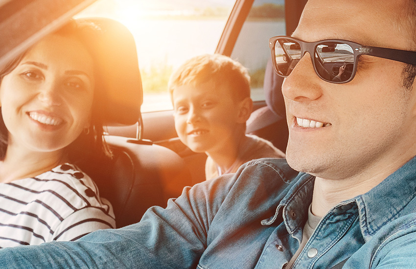 Family of three sitting in a car smiling at the camera, sunlight coming through windows
